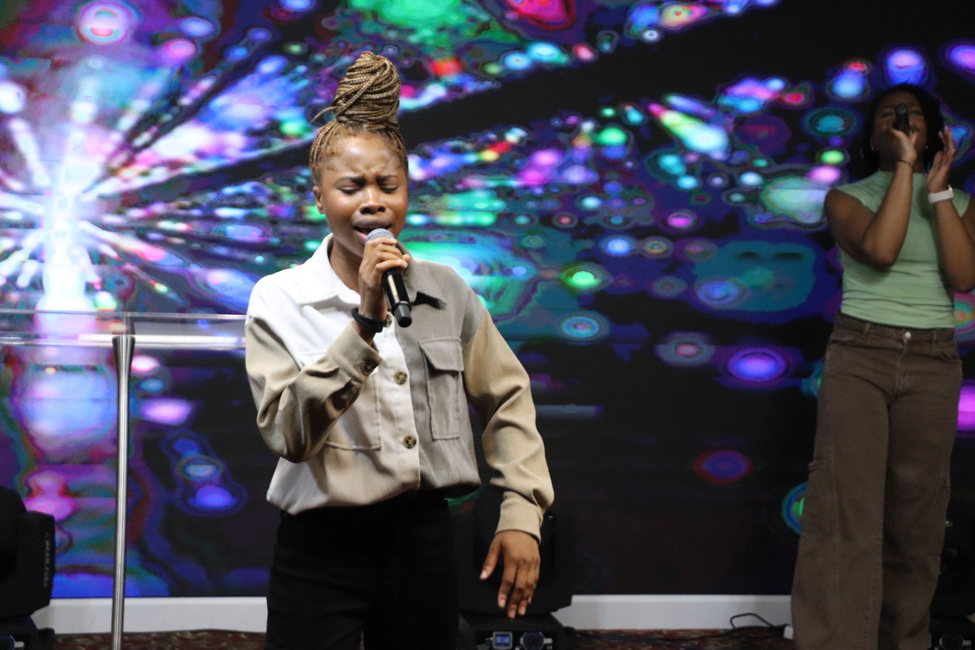 Woman singing into a microphone on stage with another singer in the background, against a colorful, blurred backdrop.