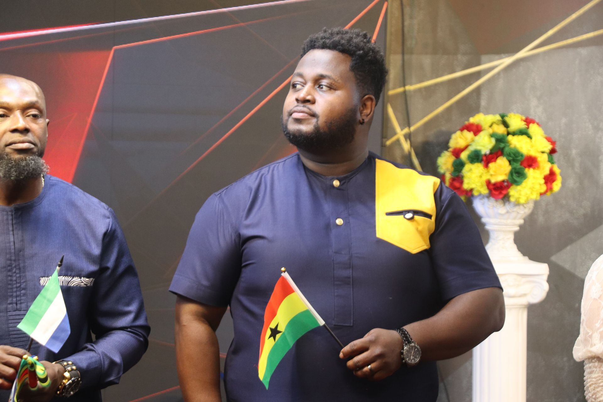 Man in blue shirt with Ghanaian flag, standing near another holding a Sierra Leone flag.