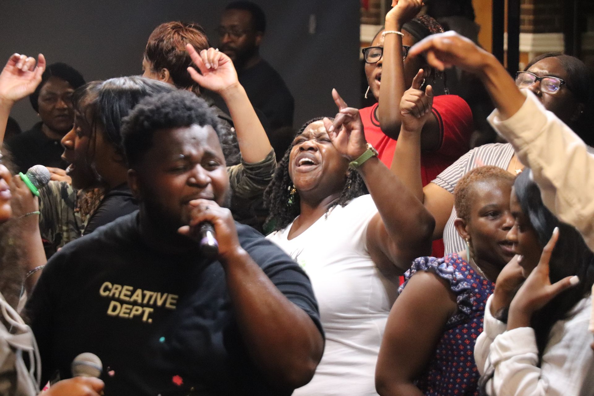 Group of people singing and raising arms, appearing joyful in a dimly lit room.