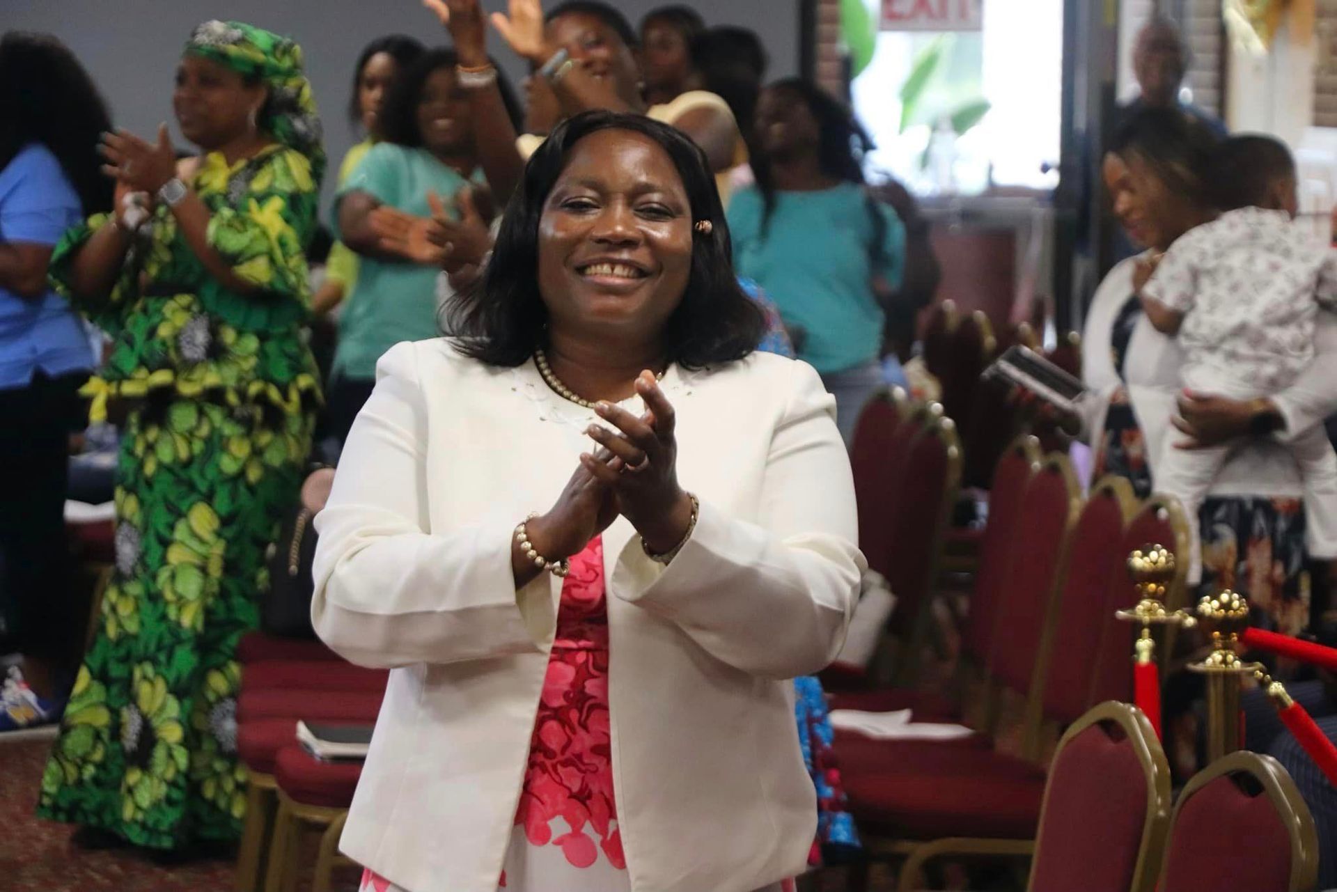 Woman in white jacket clapping, surrounded by people in a church setting.