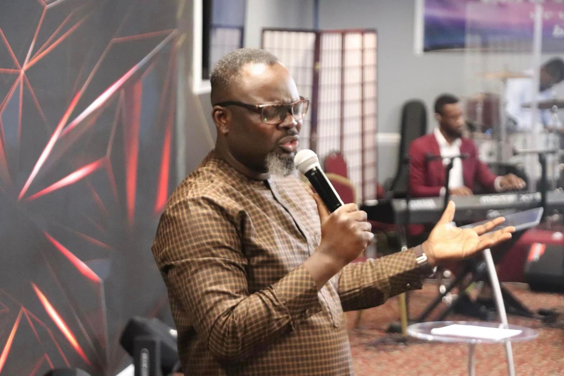 Man with graying beard speaks into microphone, gesturing. Indoors, music equipment visible.