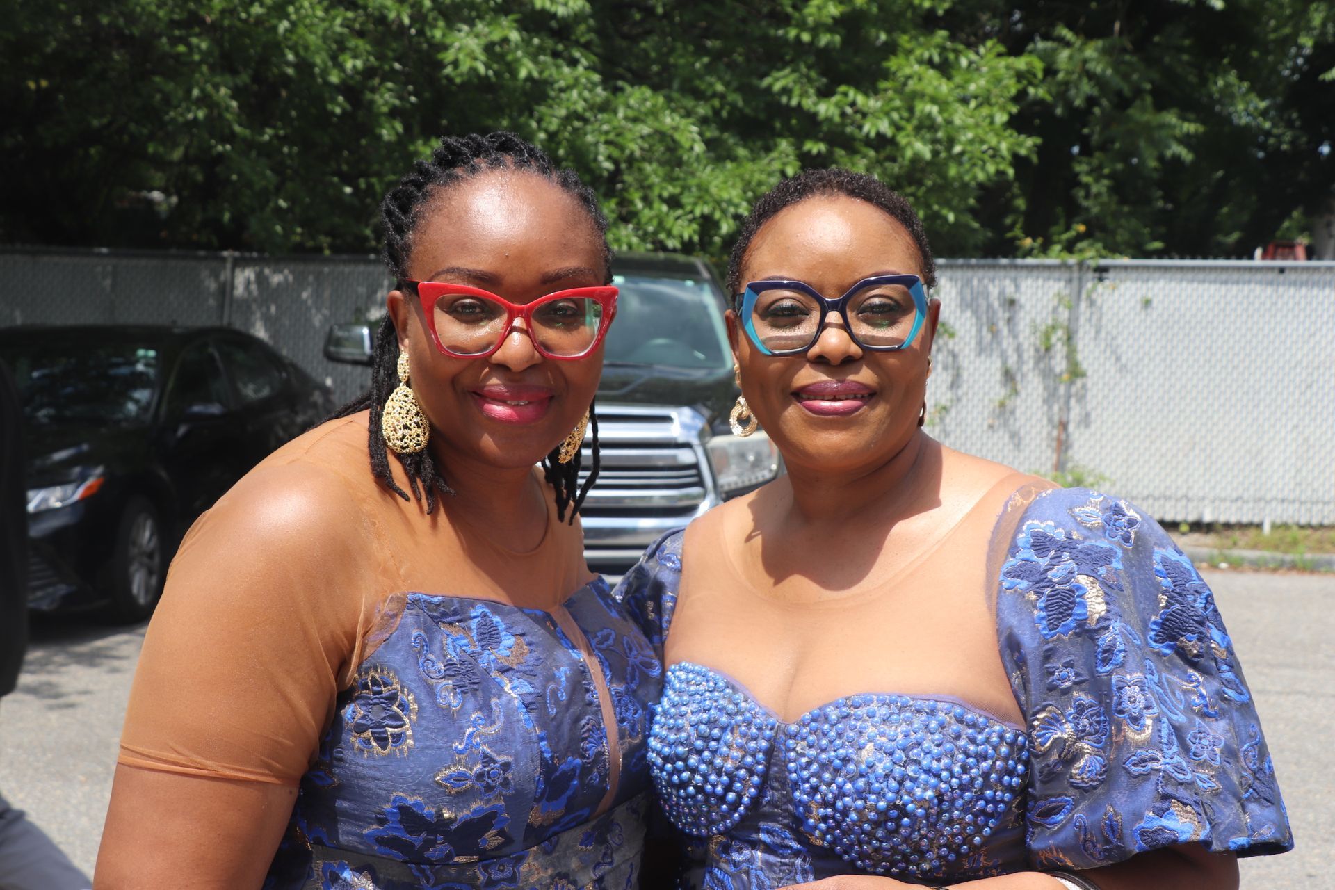 Two women in blue dresses and glasses pose outdoors.