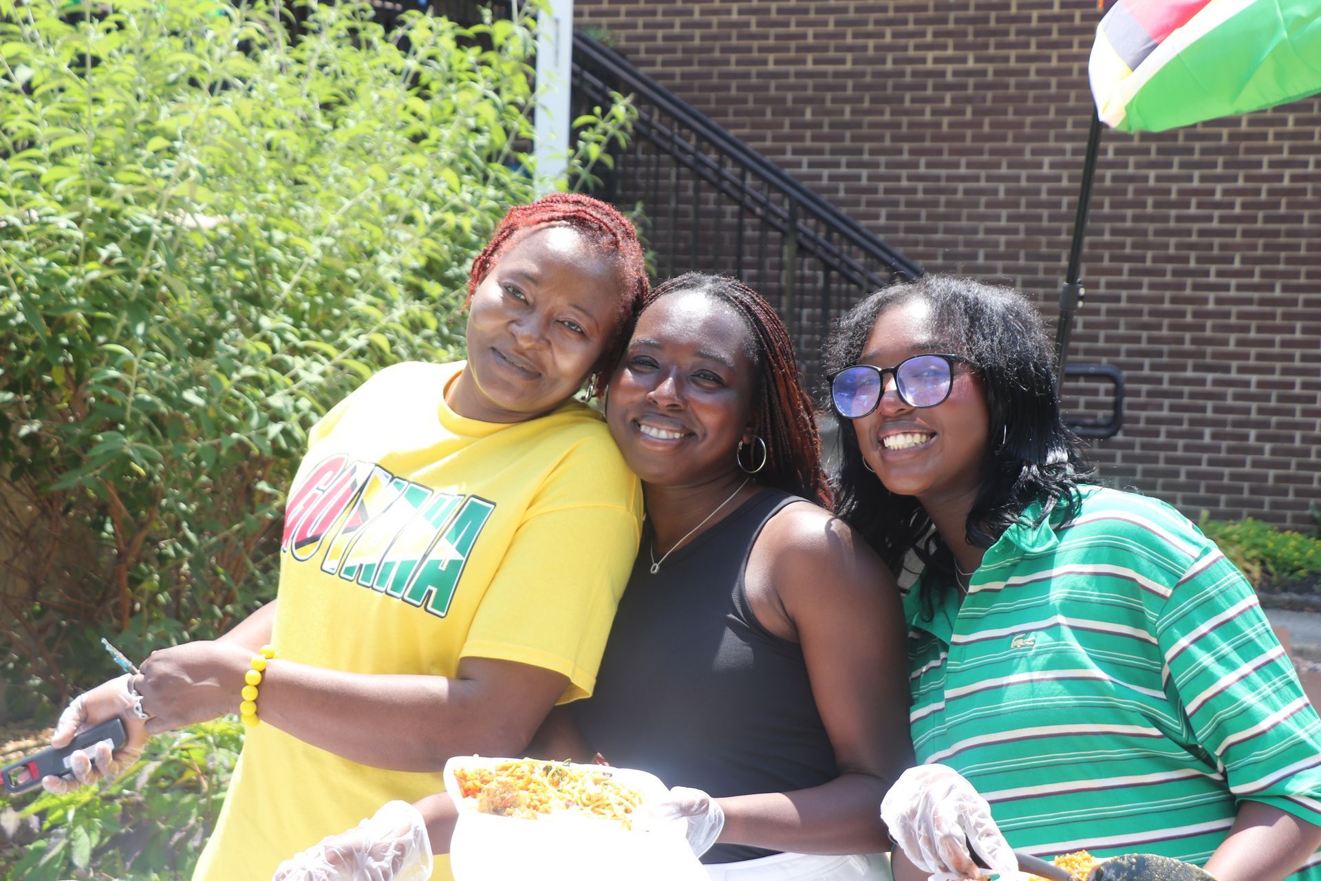 Three smiling women posing outside, one wearing a yellow shirt, one in a black tank, and the last with sunglasses.