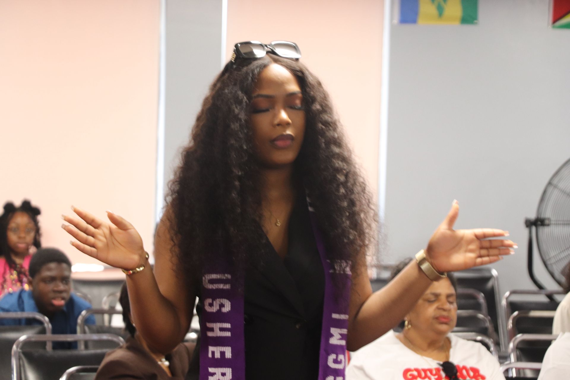 Woman with hands raised in prayer, wearing a purple sash. Indoors, other people present.