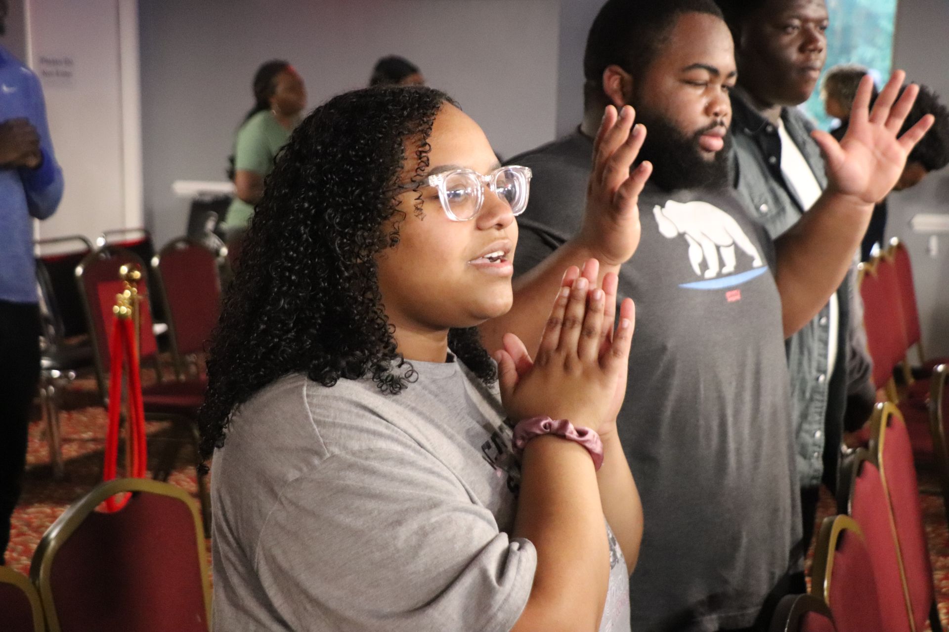 People praying with raised hands in a dim room. A woman in glasses in the foreground, focused, with others behind her.