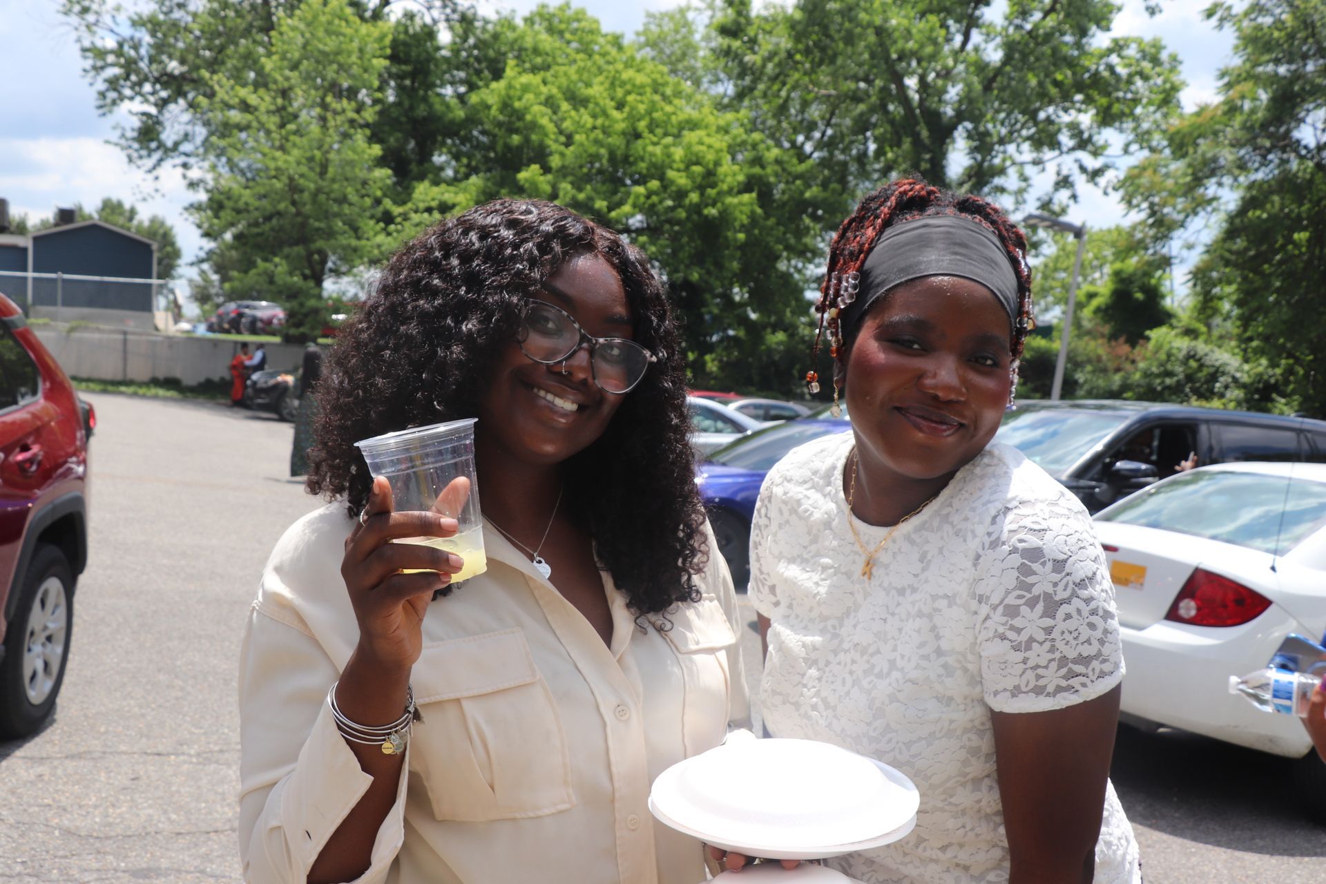 Two smiling women outdoors; one with a drink, the other with a plate. Cars and trees in background.