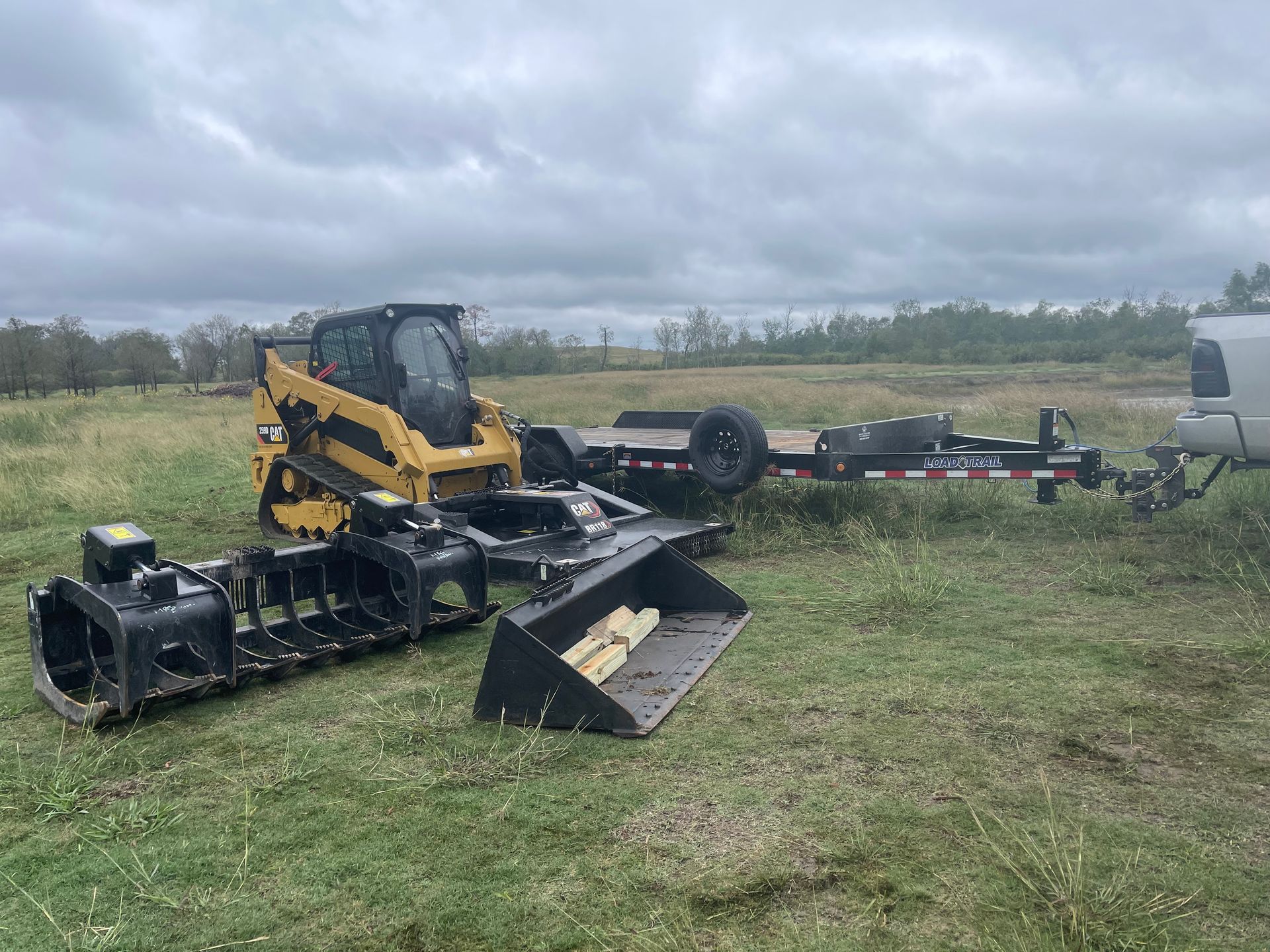 Yellow skid steer loader and attachments on a grassy field, with a trailer and cloudy sky.