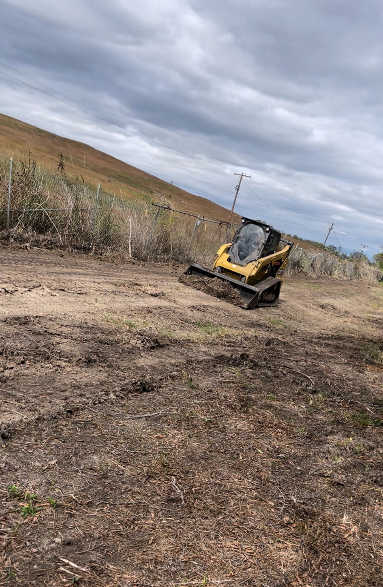 Yellow skid steer on a dirt path next to a field, against a hillside and cloudy sky.