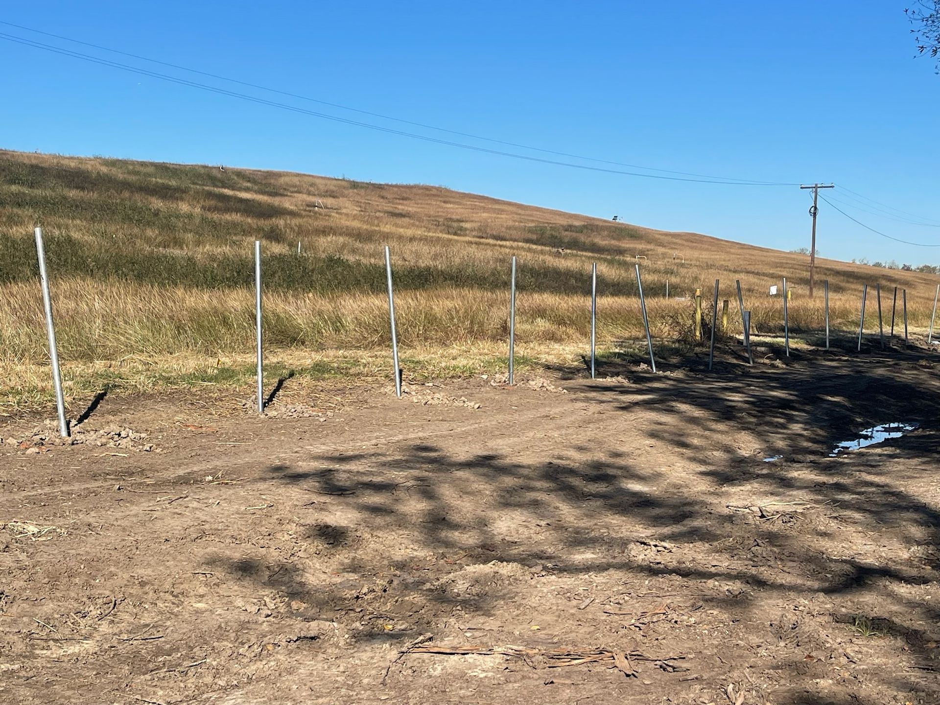 Metal fence posts set in dirt, bordering a grassy hill under a blue sky.