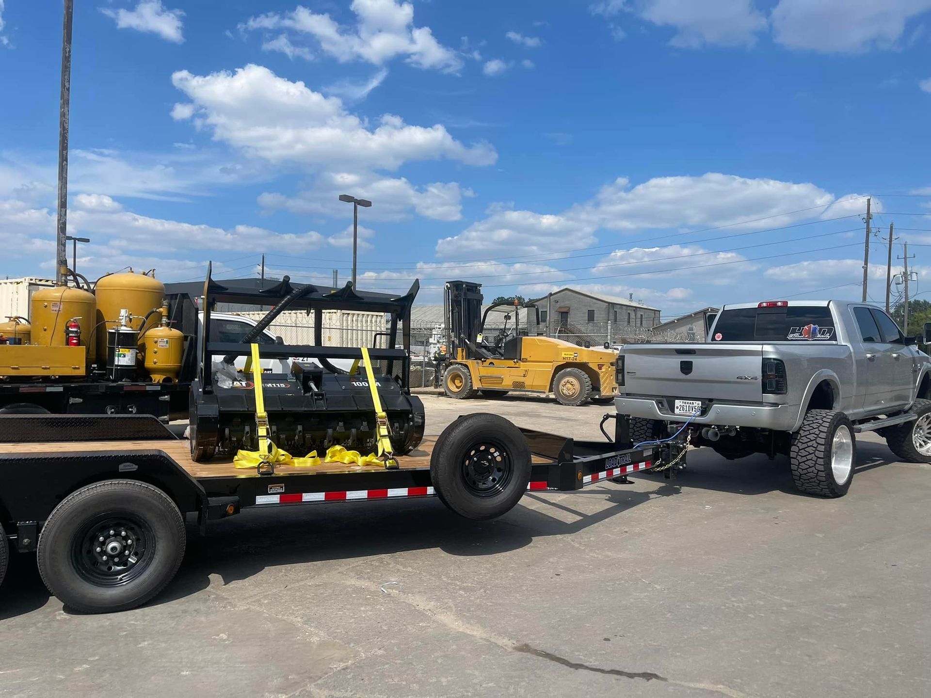 Silver pickup truck towing a trailer with construction equipment under a blue sky. A forklift is visible in the background.