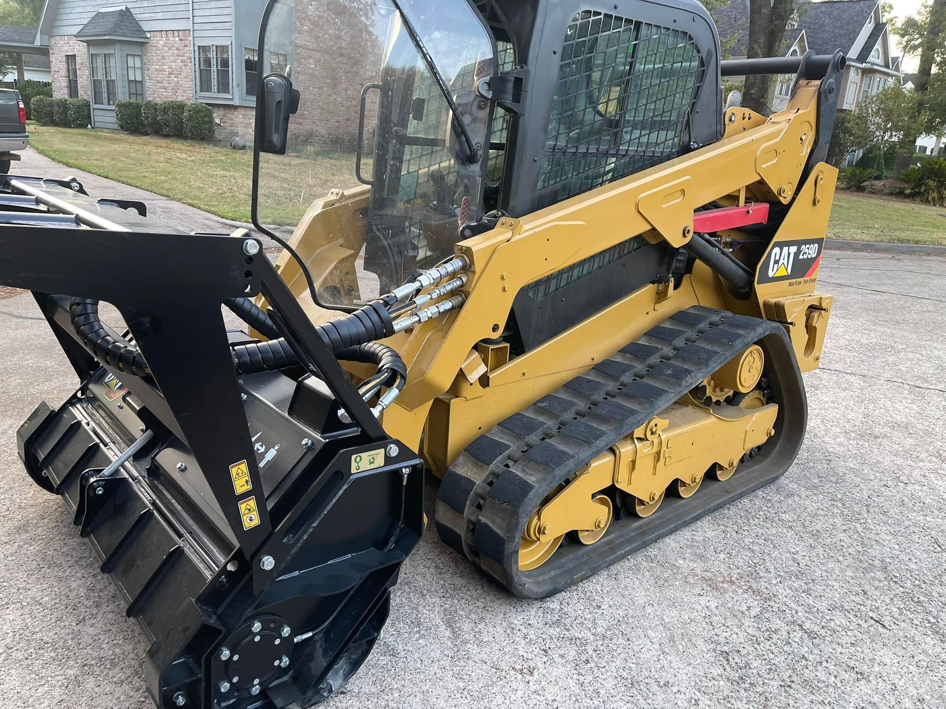 Yellow Caterpillar track skid steer with black brush cutter attachment on pavement.