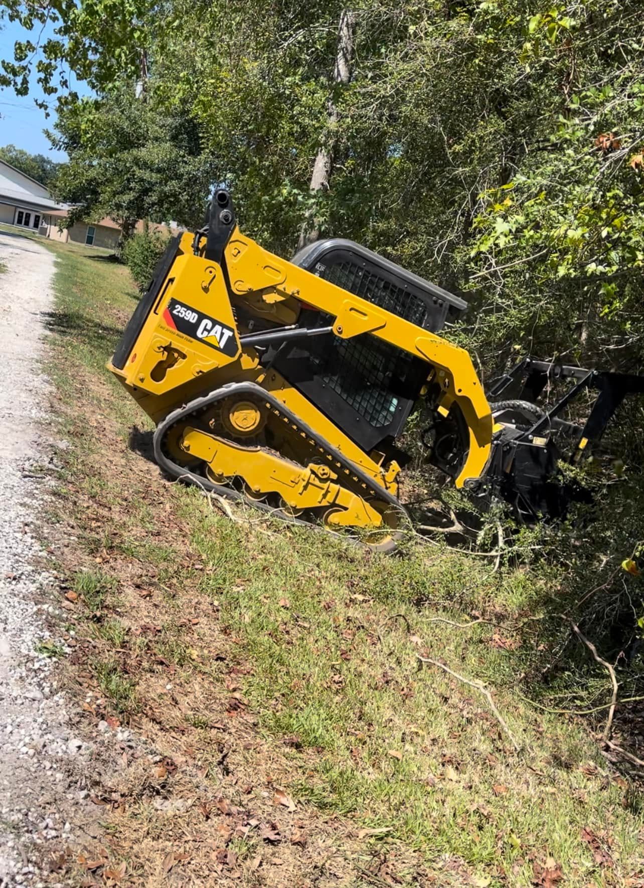 Yellow Caterpillar skid steer overturned on a grassy bank near trees.