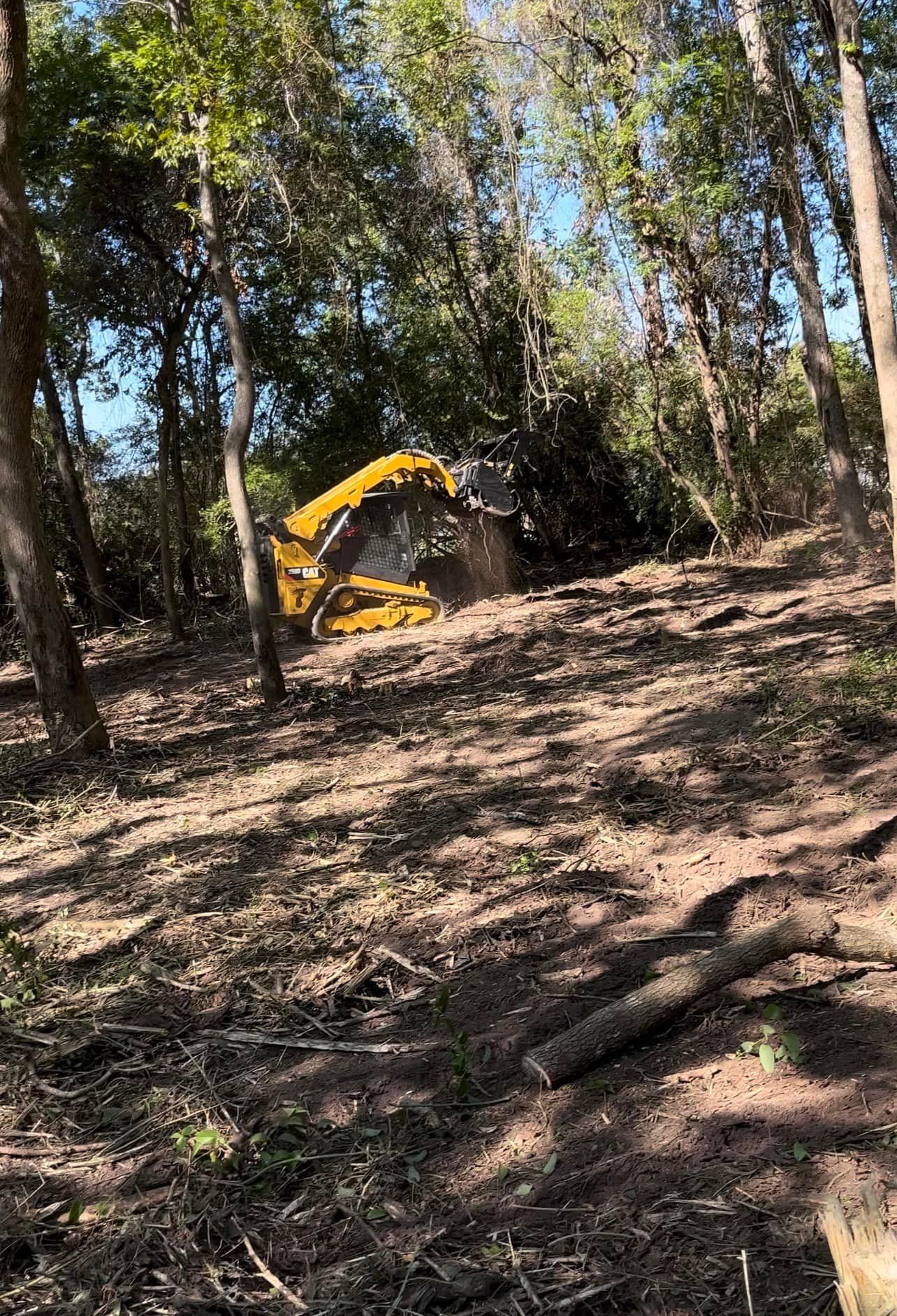 Yellow excavator clearing dirt and debris in a wooded area.
