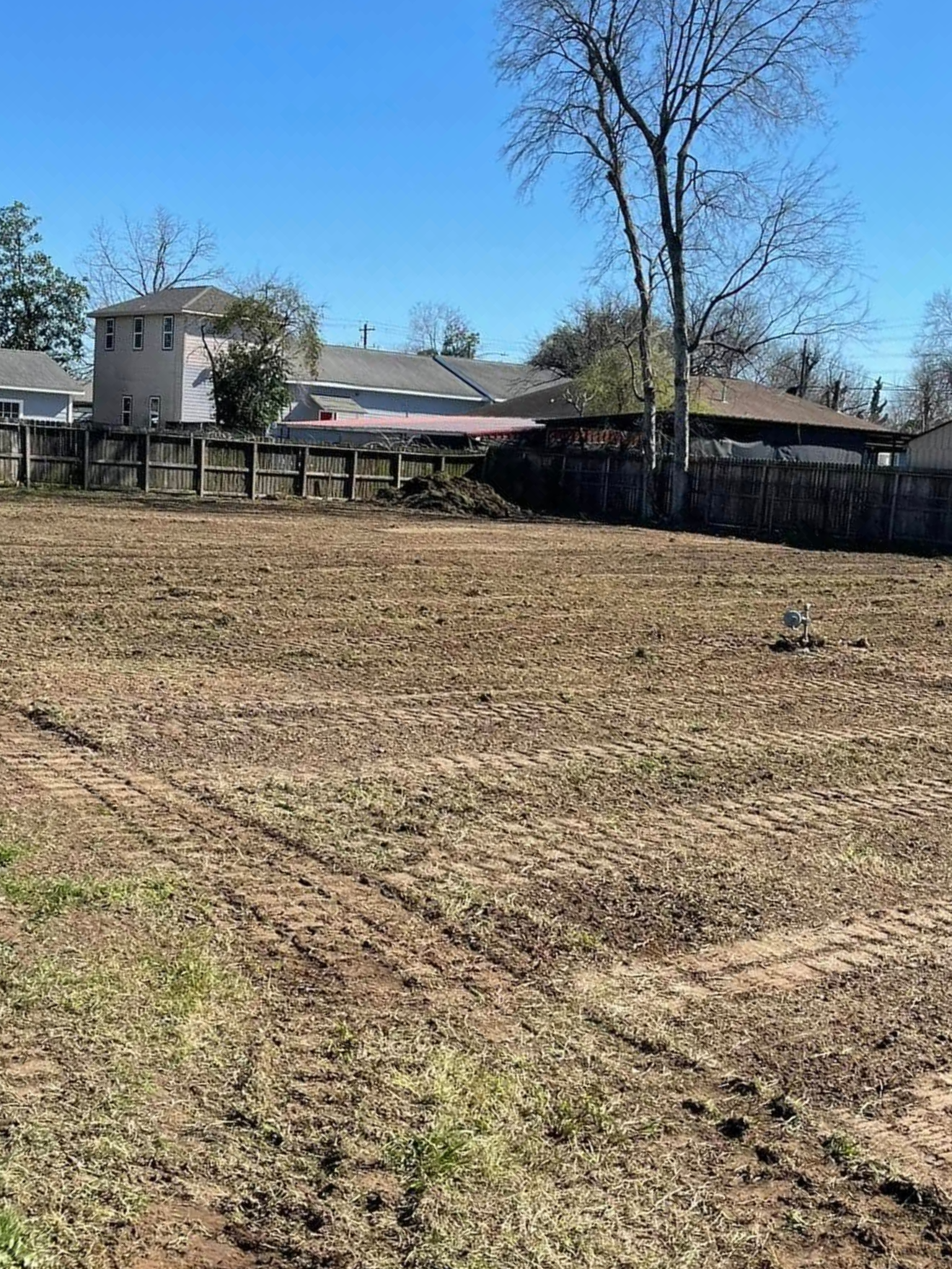Cleared dirt lot with a wooden fence and houses in the background under a blue sky.
