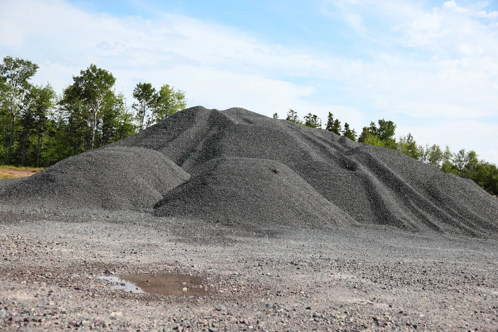 A large pile of gravel with a conveyor belt, set against a hill with colorful autumn trees.