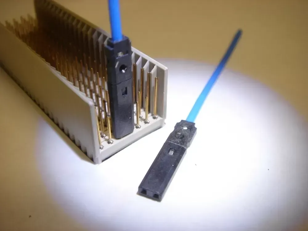 A blue-wired jumper connector being plugged into a multi-pin electronic header block next to a detached matching connector.