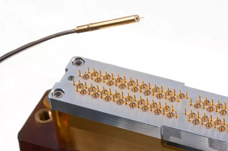 A metallic probe cable positioned above a rectangular component featuring an array of gold-colored spring-loaded pins.