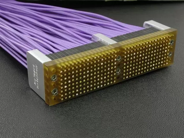 A high-density rectangular electrical connector with many small gold pins and a bundle of purple wires attached to the back.