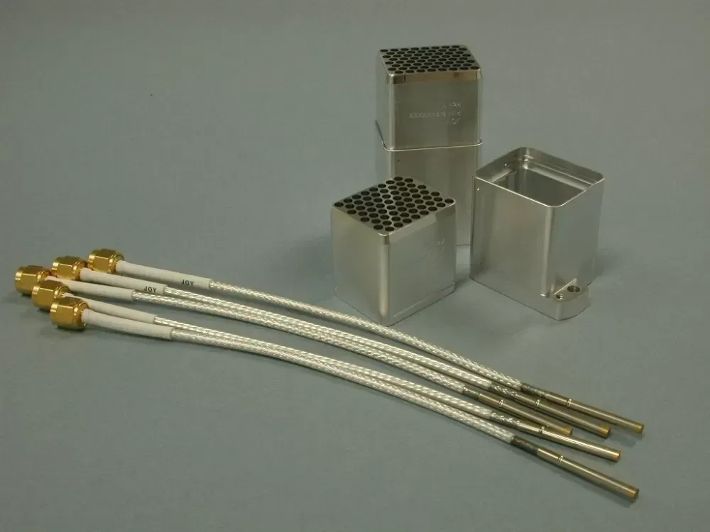 Four RF cables with gold connectors next to two metal housings, one with a grid of small holes, on a gray background.