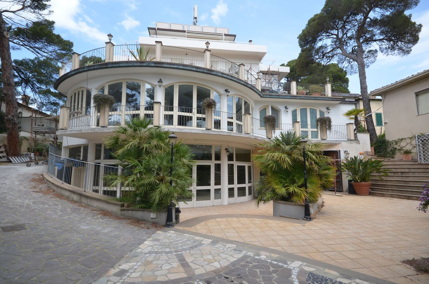 Villa bianca dalle forme sinuose con ampie finestre, cortile in pietra e alberi sotto un cielo azzurro.