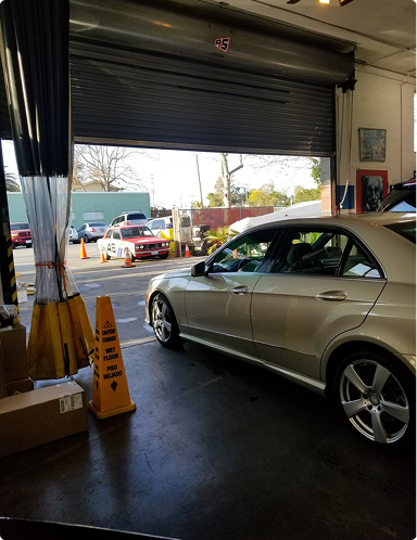 Silver sedan inside a garage with a partially open roll-up door, looking out to a street scene. | Hudson Automotive