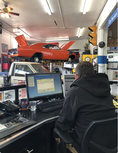 Person at a computer inside an auto repair shop with a red car lifted in the background | Hudson Automotive