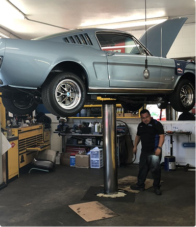 Classic car lifted on a garage hoist, with a mechanic standing below in a workshop. | Hudson Automotive