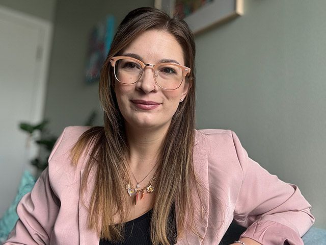 A woman wearing glasses and a pink jacket is sitting on a couch.
