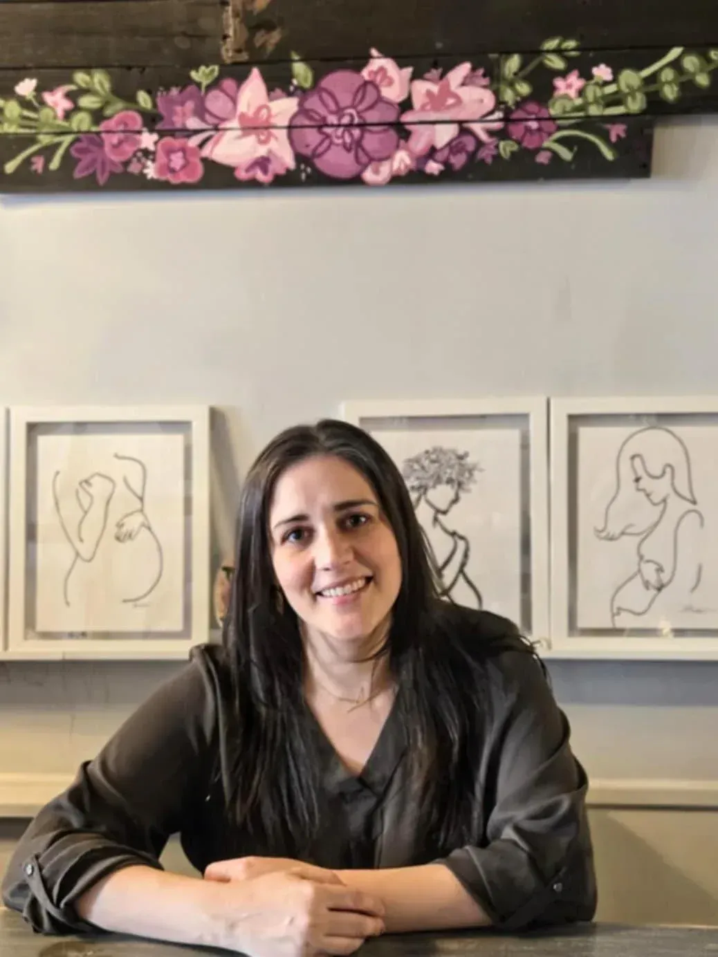 Kari Tetzlaff Kari Tetzlaff, owner of Rosy Glow Maternity is sitting at a table in front of a painting on a wall.