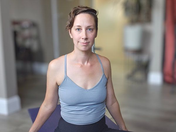 Woman in a gray tank top sits on a yoga mat, looking forward. Indoor setting, soft lighting.