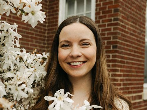 Woman smiles near blooming white flowers and brick building.