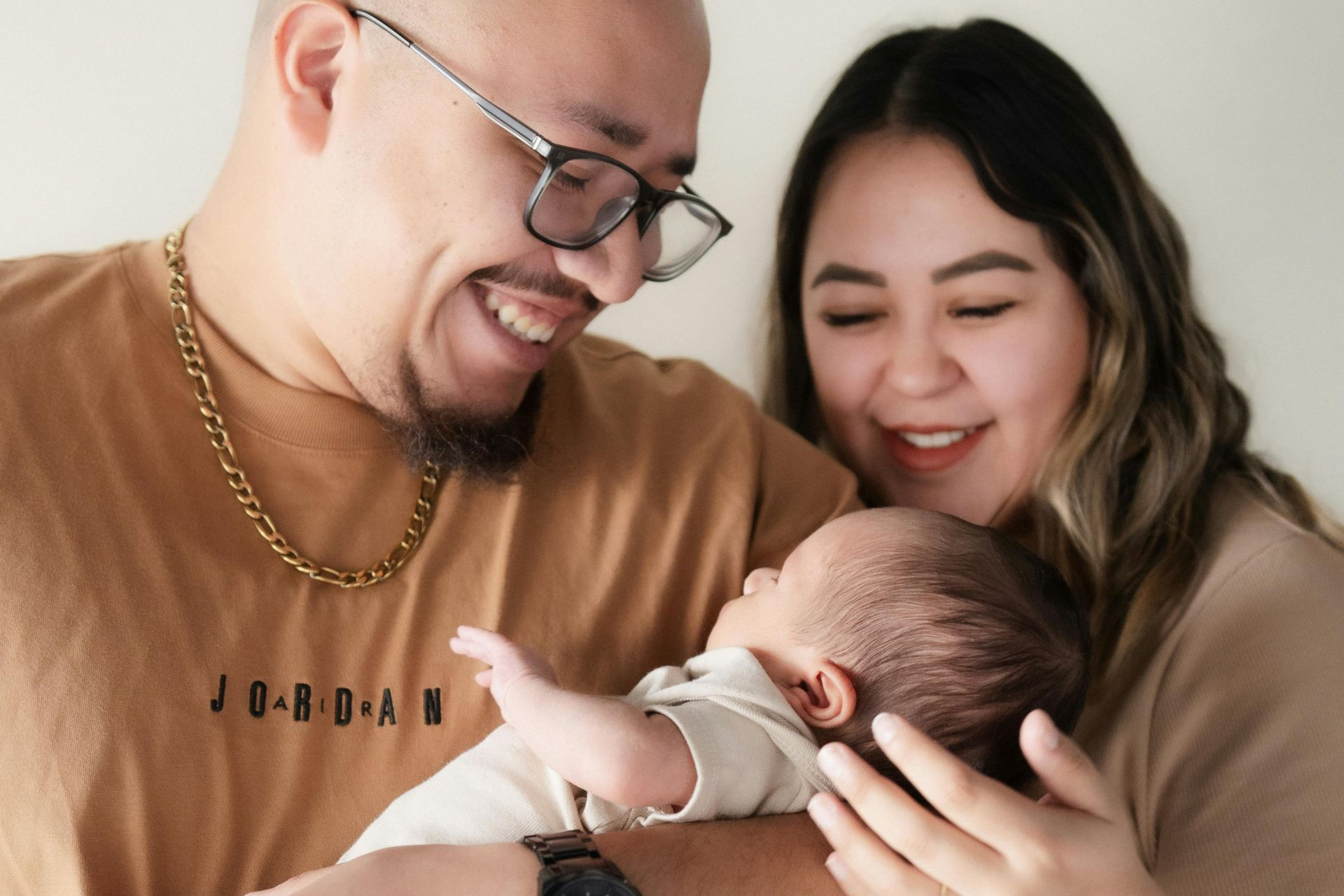 Parents smiling at newborn baby. Man in glasses, gold chain, tan shirt. Woman in tan top. Neutral background.
