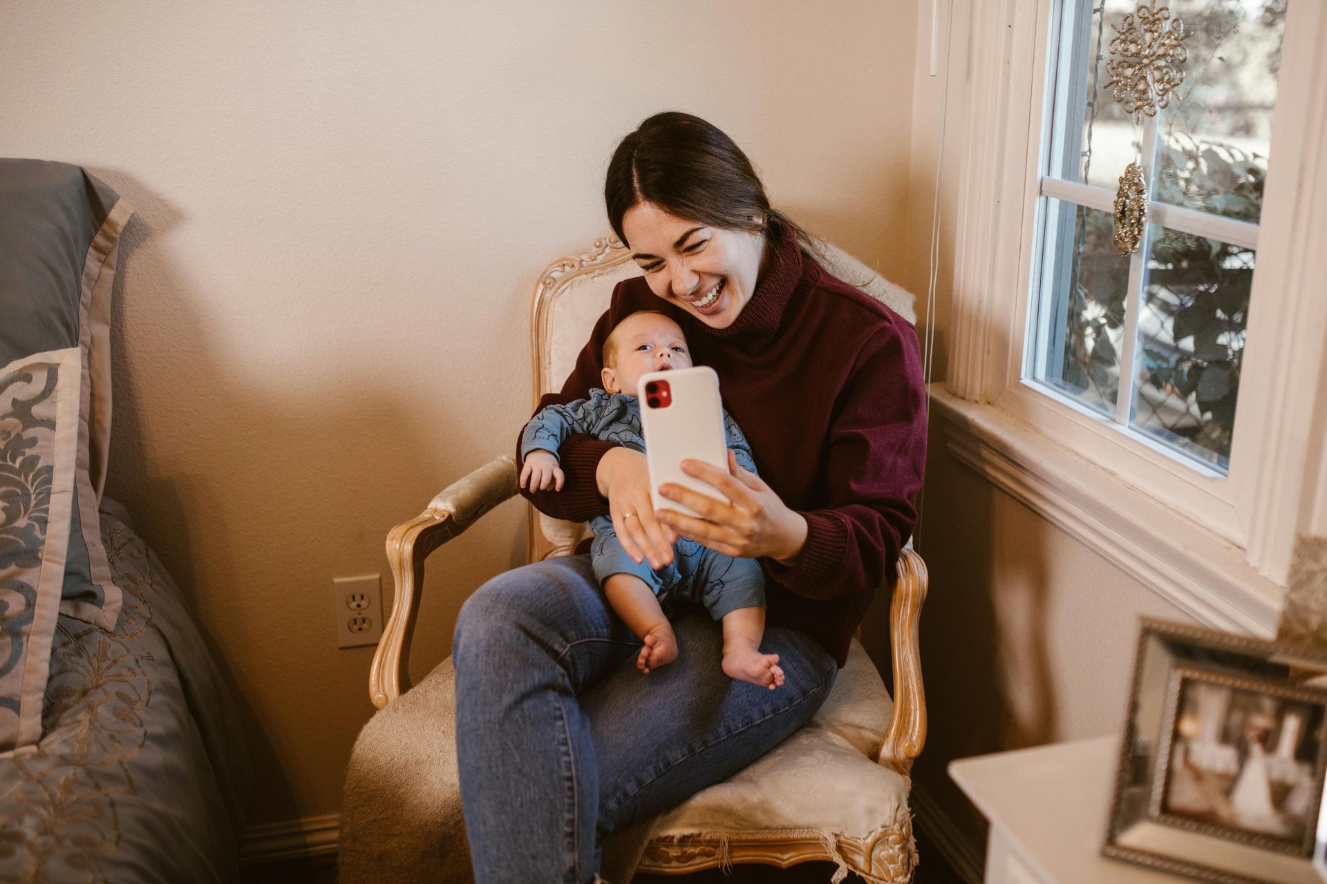 Woman in burgundy shirt holds baby, taking a selfie on a phone, by a window.