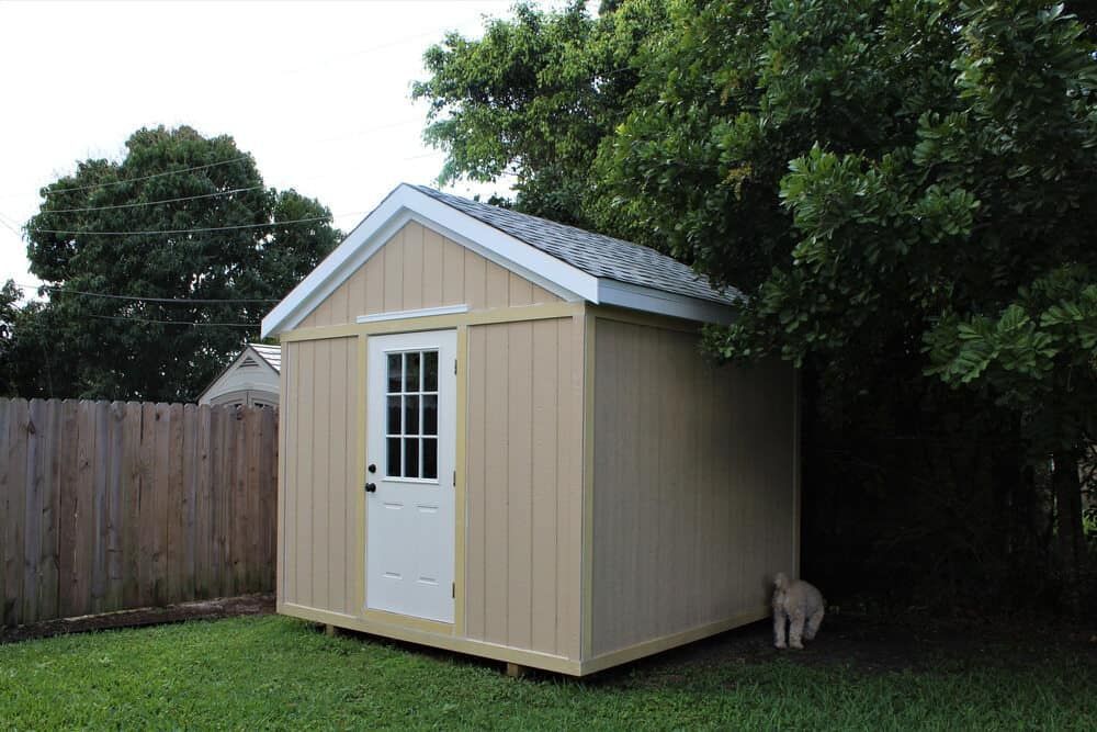Tan Shed With White Door, Set on Green Grass, Next to a Wooden Fence and Tree — Clearspan Builders In Royalla, NSW