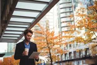 Man in suit, holding coffee and tablet, walking under a modern canopy, autumn trees in background.