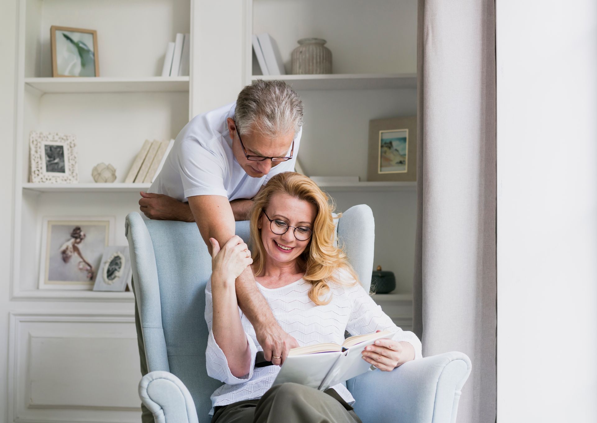 Man leans over woman sitting in armchair, both reading a book near a window and bookshelf.