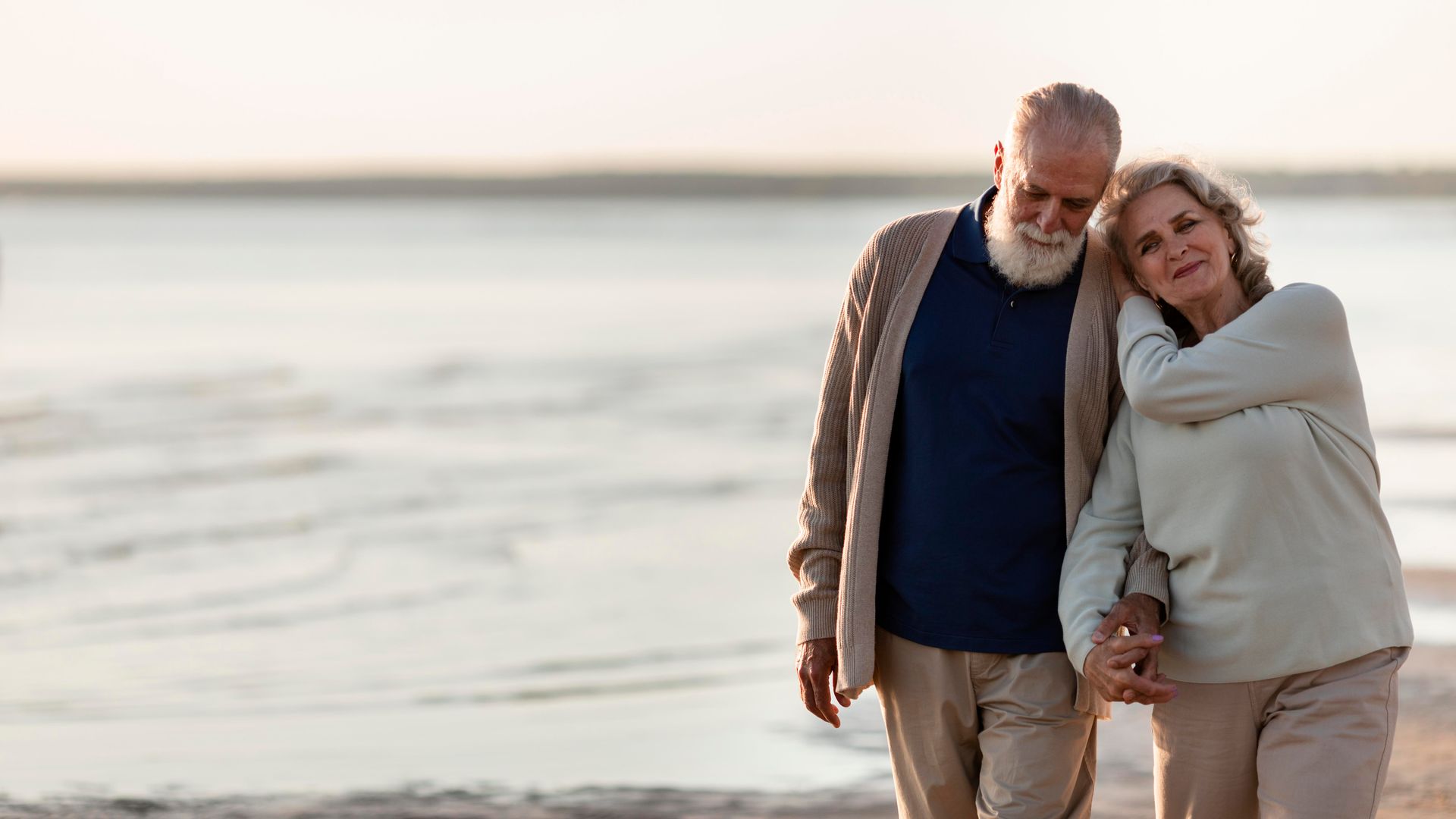 Elderly couple walking, holding hands, on a beach. One leans on the other, smiling.