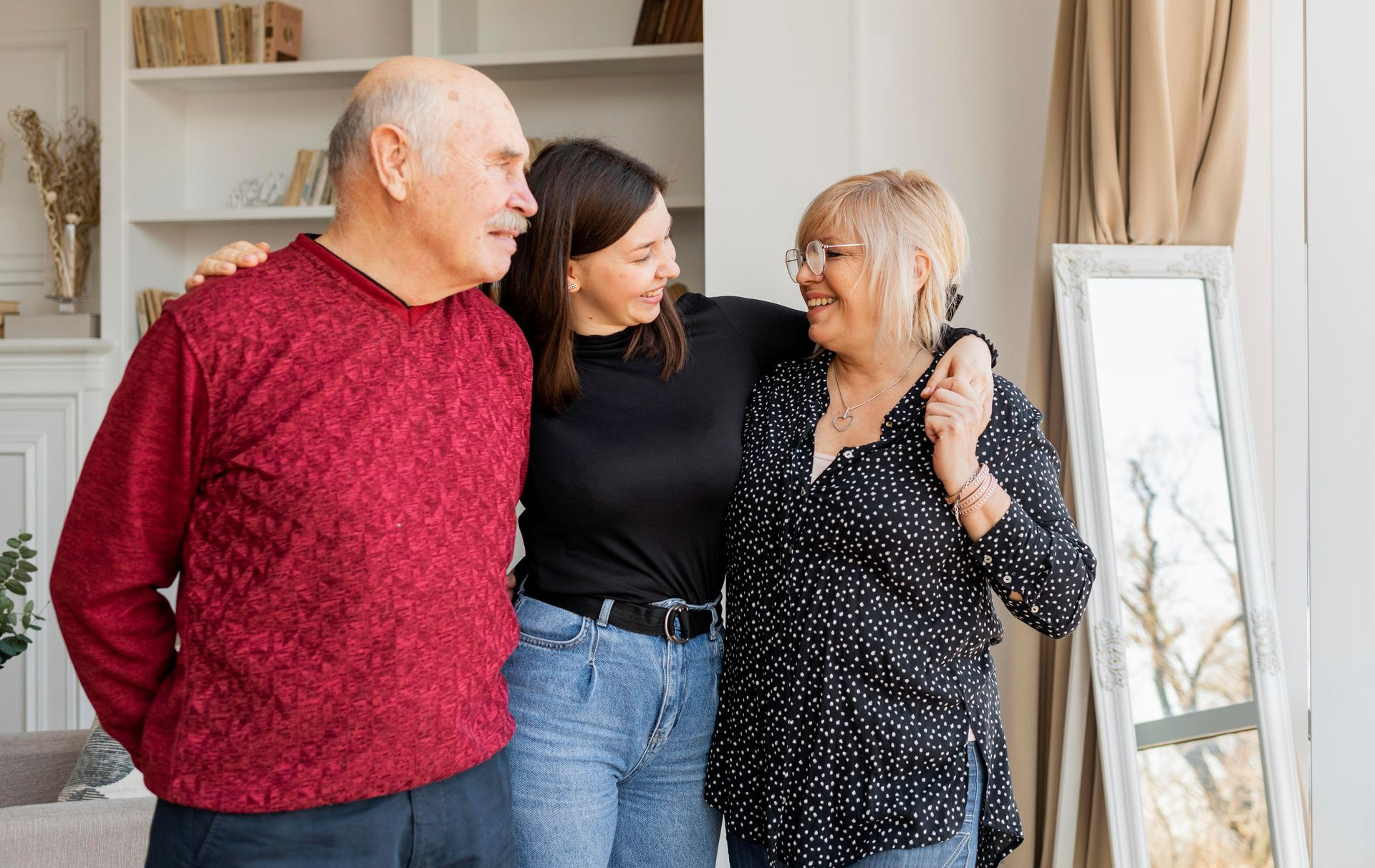 A woman embraces two older adults indoors. The group smiles. The man wears red, the woman has glasses.