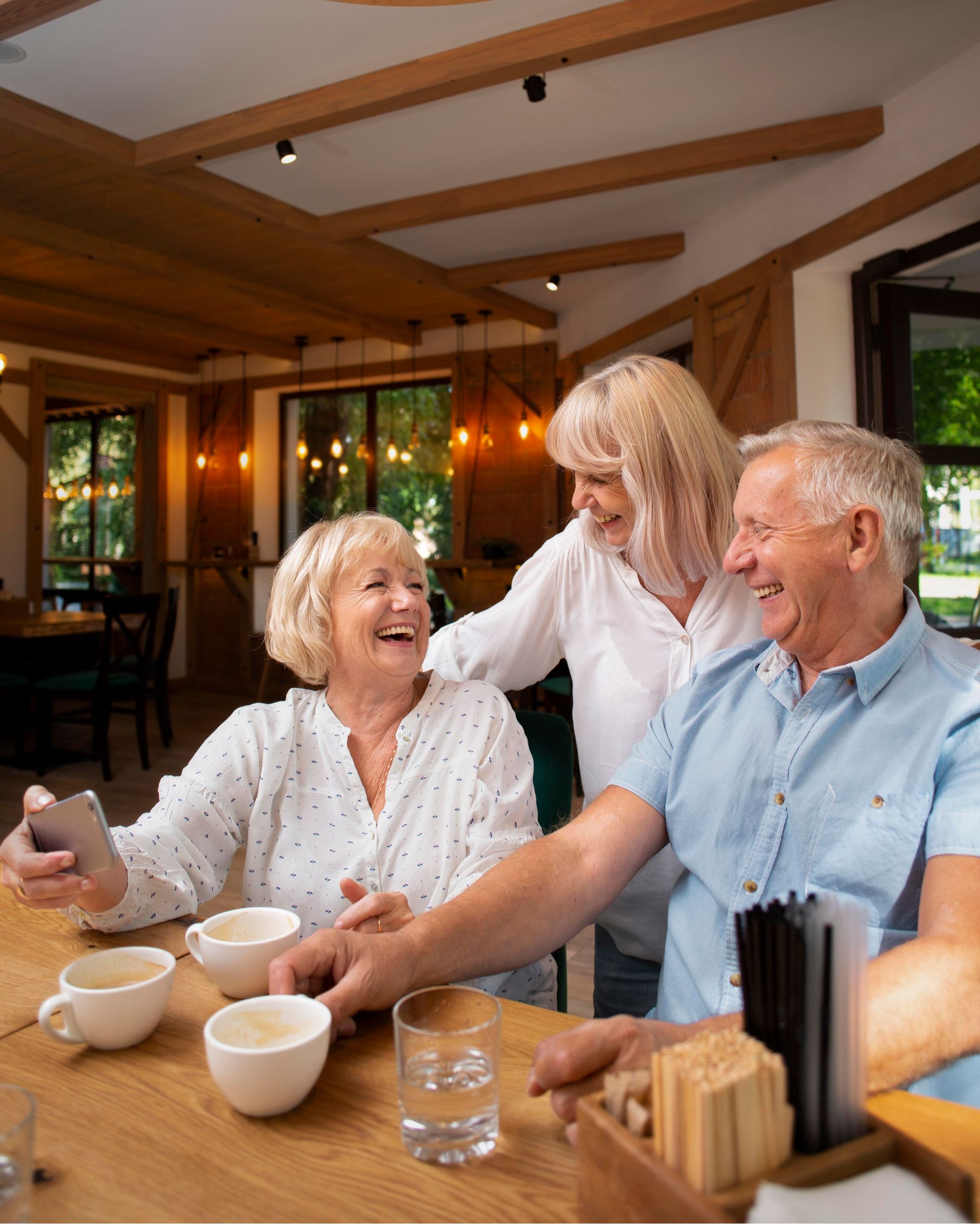 Three people laughing together at a cafe counter. One holds a phone, two white coffee cups on the table.