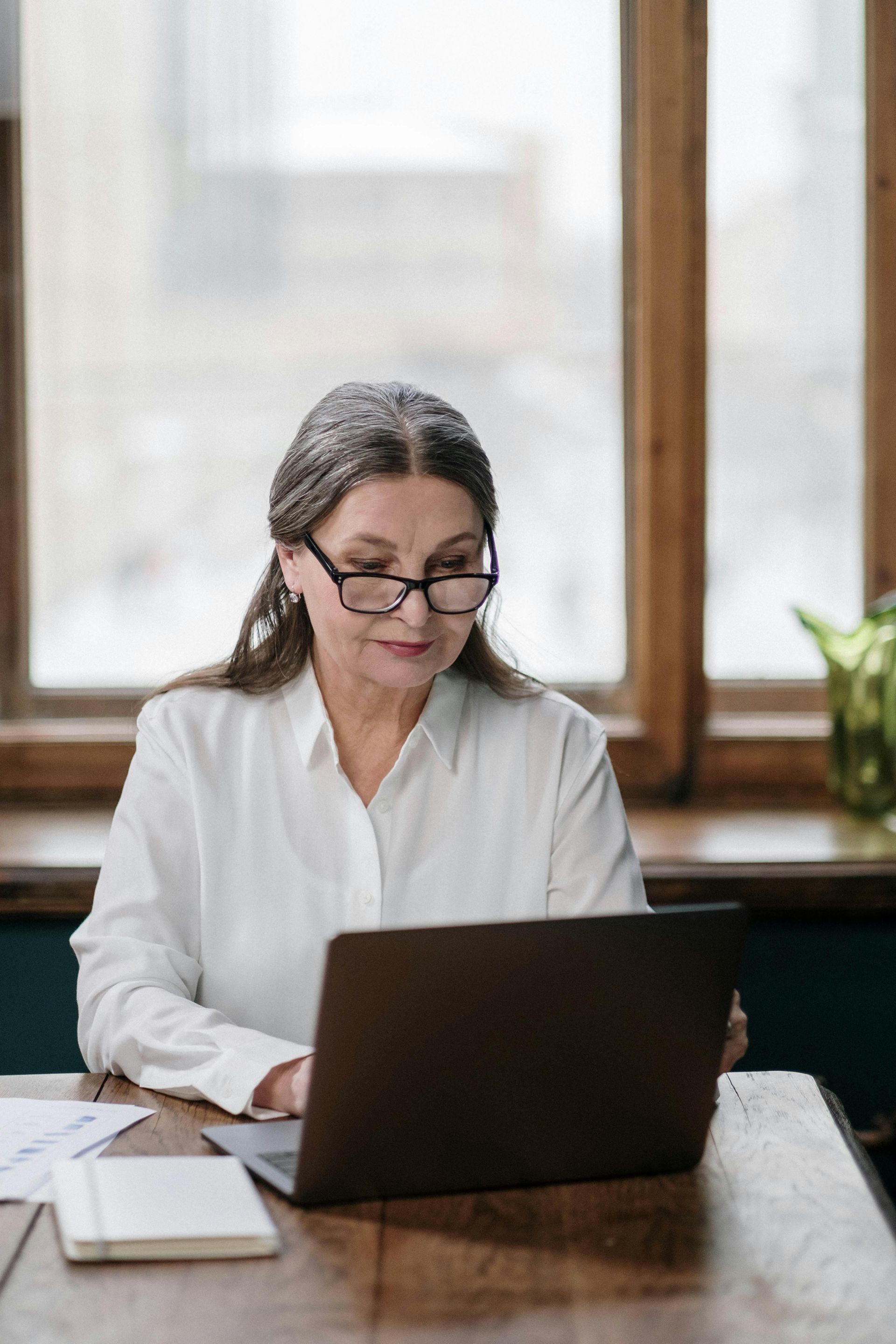 Woman with gray hair wearing glasses, looking at a laptop at a wooden table near a window.