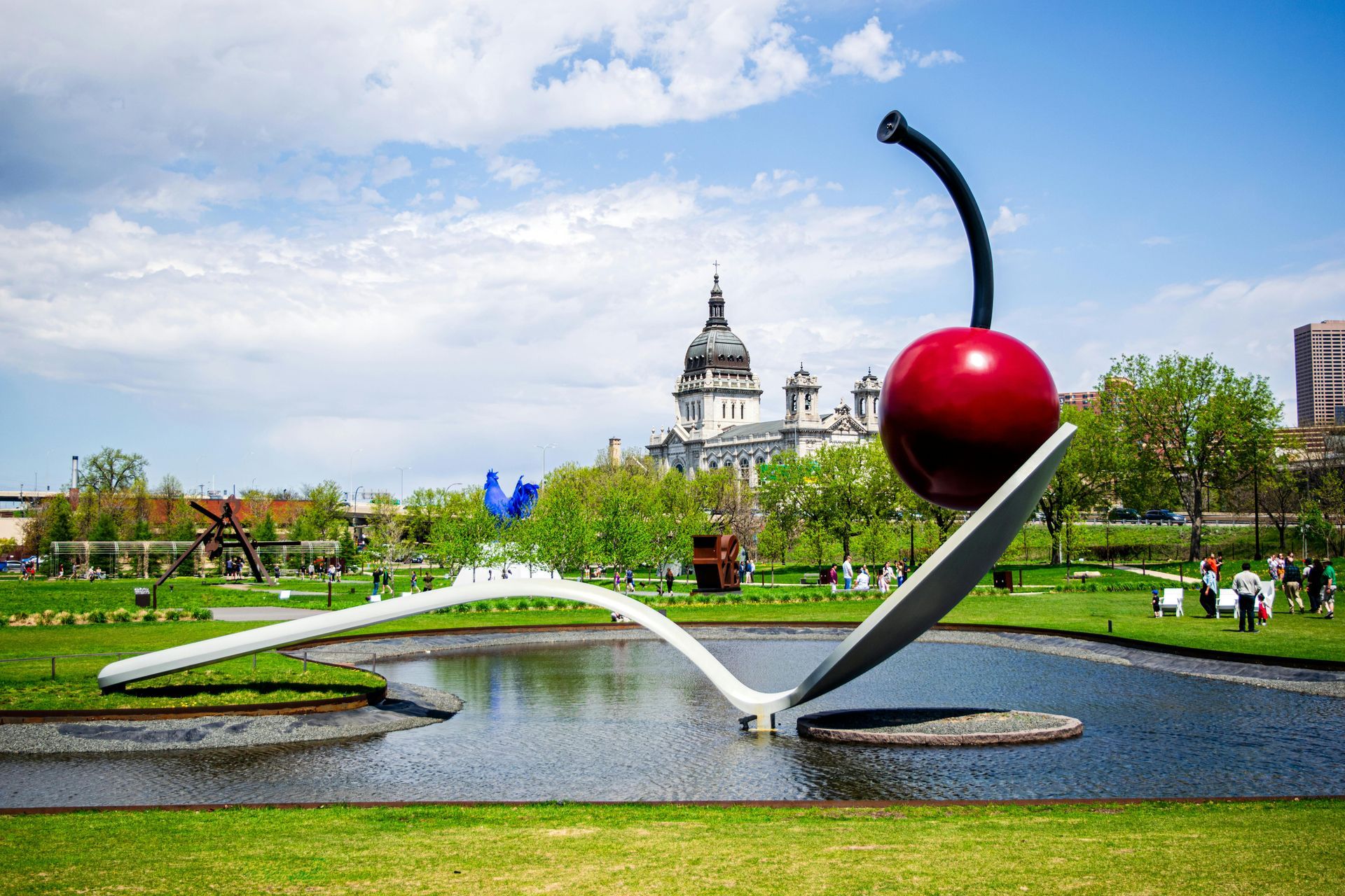 Giant spoon and cherry sculpture in a pond, with a cityscape and sunny sky in the background.