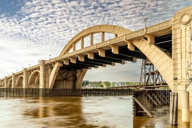 Arch bridge spanning a river, beige concrete with arched supports, cloudy sky.