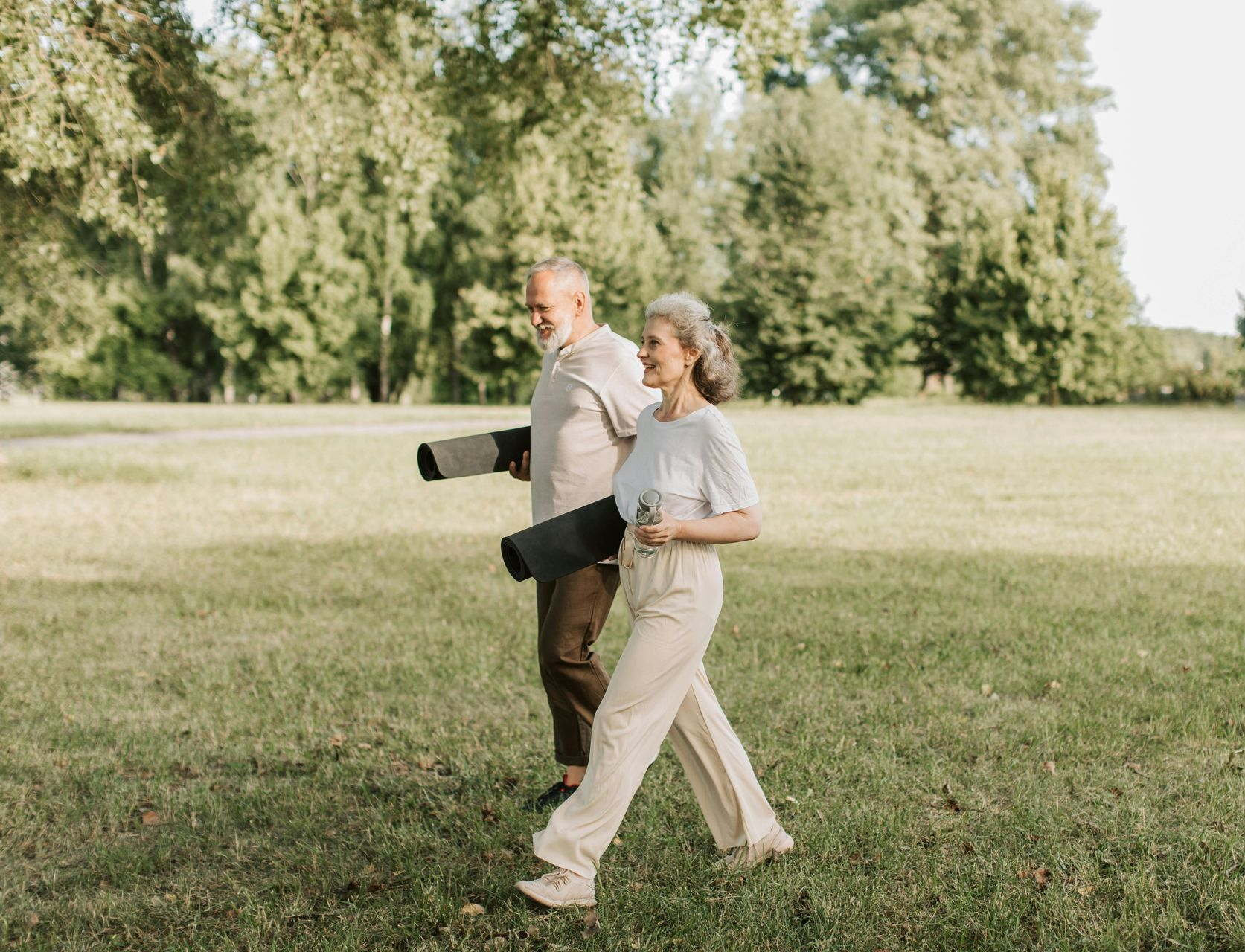 Couple walking through a grassy park holding yoga mats, trees in the background.