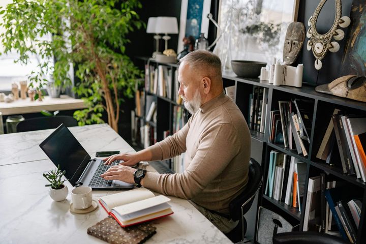 Man typing on a laptop at a white table in a room with a bookshelf.