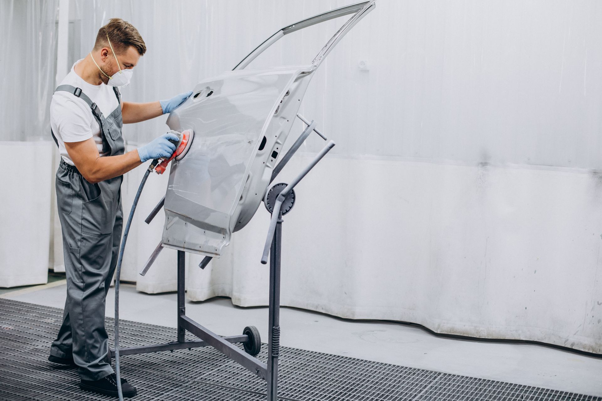 Man in overalls and mask sanding a car door in a workshop.