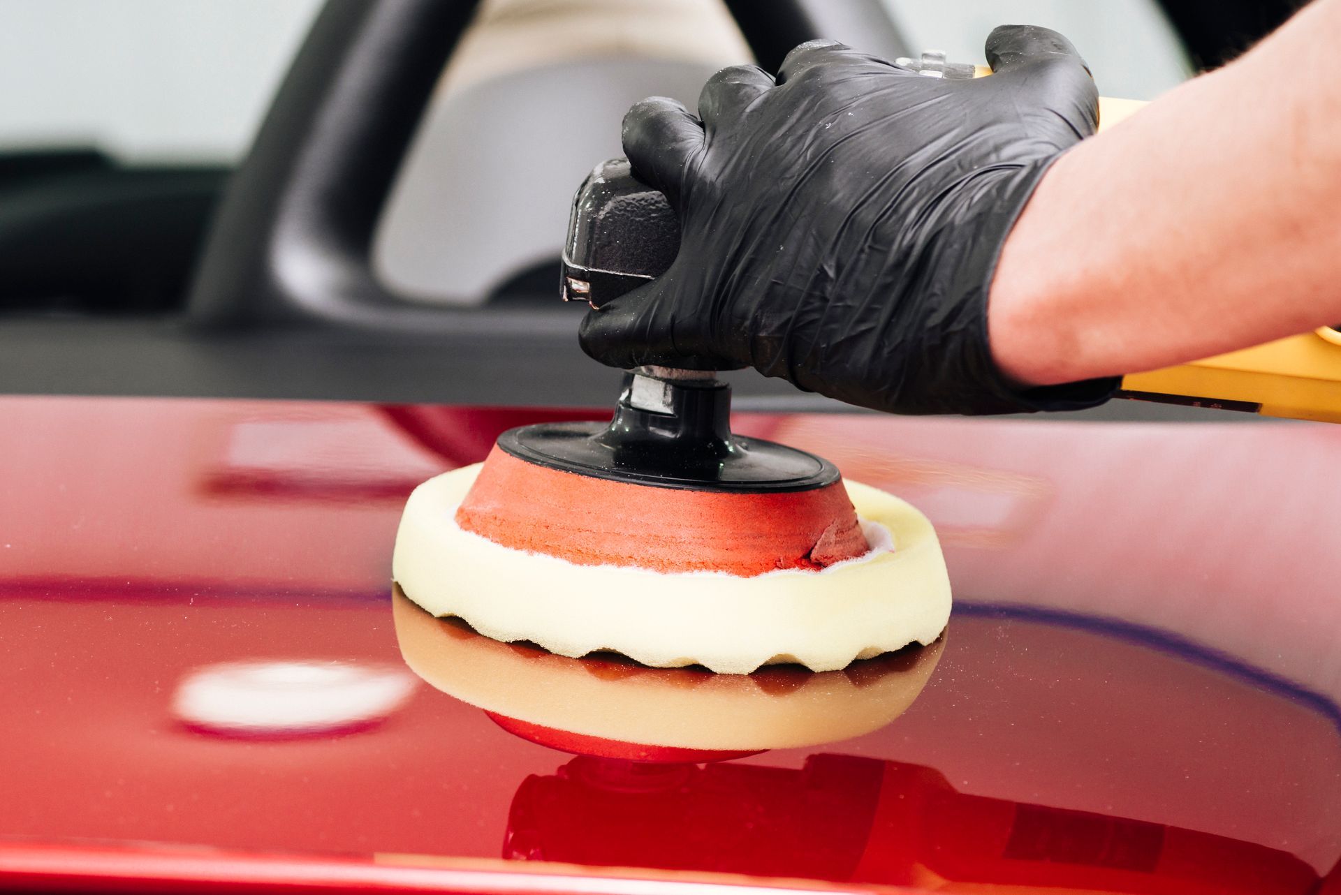 Hand in black glove polishing red car with an electric buffer, in a garage setting.