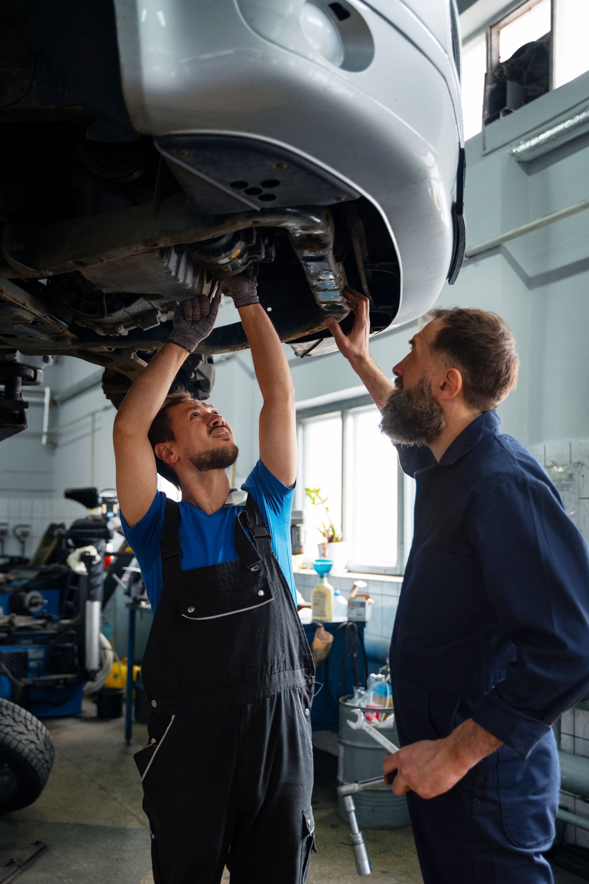 Two mechanics working under a car in a garage. One points, the other reaches up with tools.