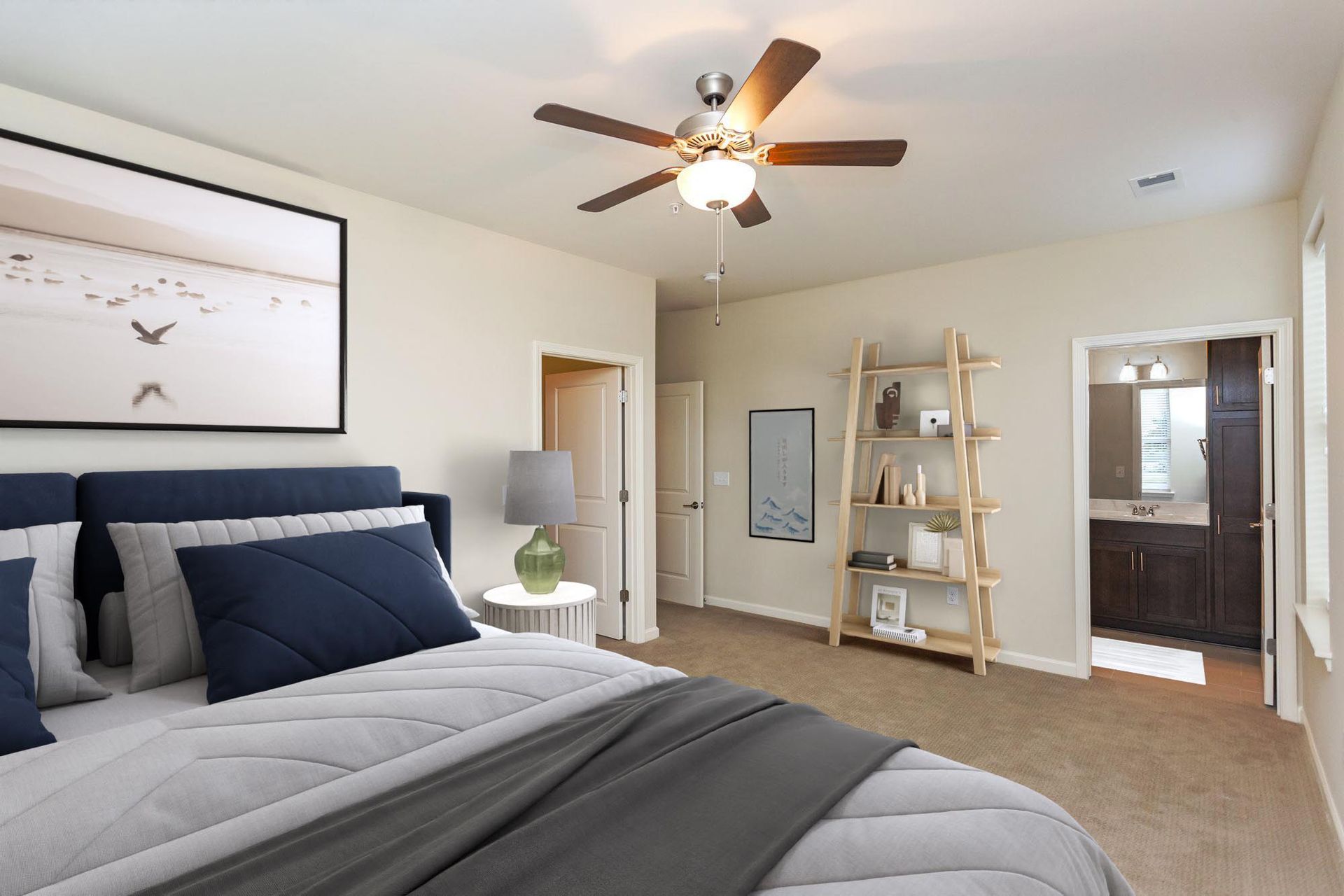 A bedroom with a bed featuring blue pillows, a ceiling fan, a wooden bookshelf, and an open doorway leading to a bathroom.
