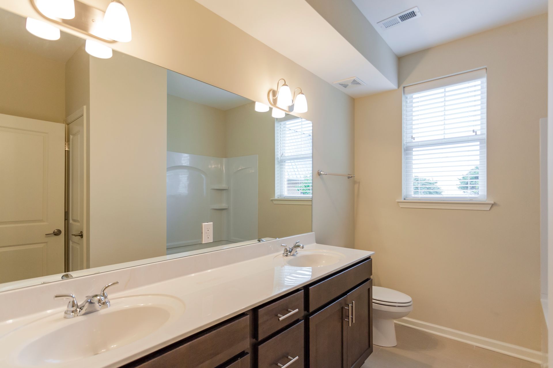 A bright bathroom features a long vanity with a sink, large mirror, wooden cabinets, and a window next to a toilet.