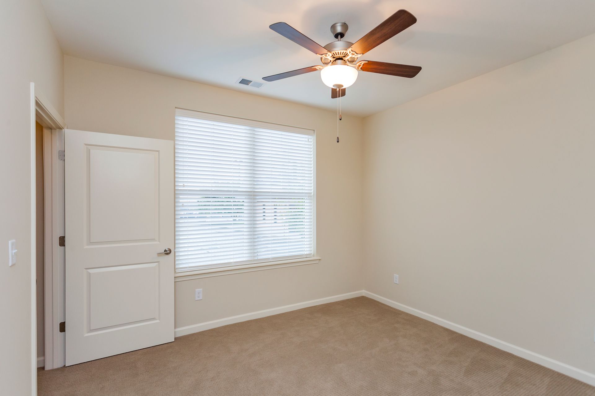 Empty room with beige walls, tan carpet, a closed white door, and a ceiling fan over a window with closed blinds.
